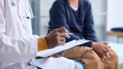 A doctor holds a clipboard and a pen and a young child sits on an examination table.