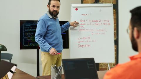 Middle aged man teaching programming concepts on whiteboard to adult male student in classroom setting, discussing code structure with laptops and digital screen visible.
