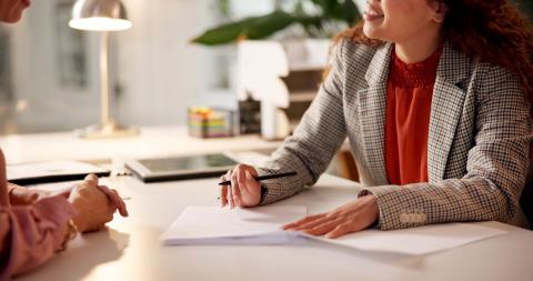 Two people sit across a desk from each other. One holds a pen and there are papers on the desk.
