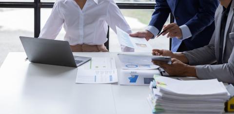 Three people gather around a table, looking at a laptop and pages printed with graphs and charts.