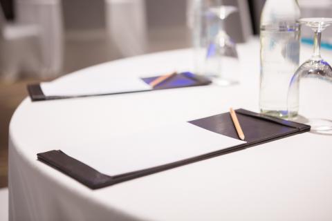 A notepad, pen, and empty glass are set on a table covered in a white tablecloth.