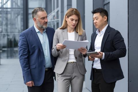 Three adults stand outside an office building wearing suits and looking at a set of papers.